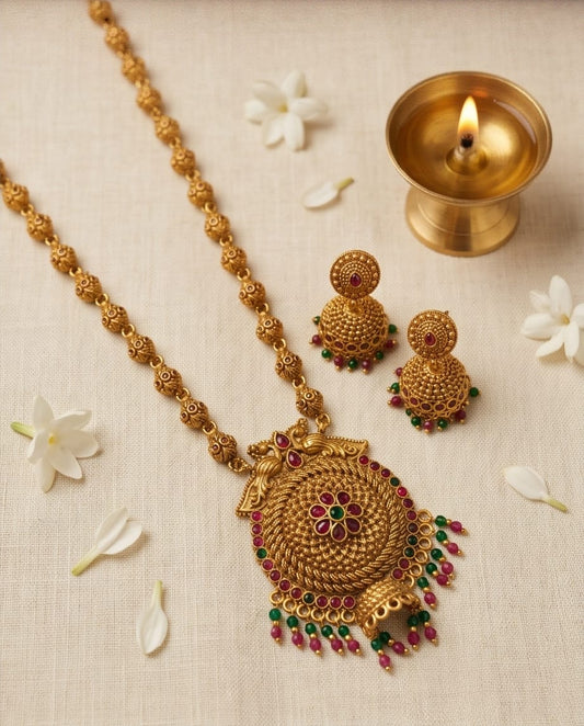 Gold necklace with intricate design and gemstones on a beige background with flowers and a small lamp.