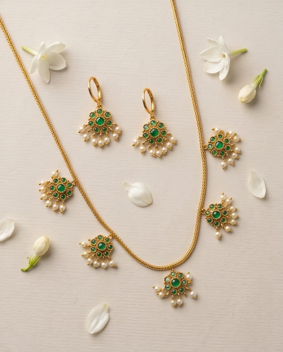 Gold necklace with green gemstone and pearl accents on a light background with white flowers.